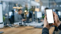 Male hand holding smartphone with blank white screen in modern office environment with blurred colleagues and workspace in background.