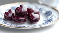 Heart-shaped chocolate macarons with glossy burgundy shells and white pearl decorations arranged on elegant porcelain plate for romantic occasions.