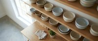 Handmade ceramic bowls displayed on wooden shelves near window with natural light. Artisan pottery collection in neutral earth tones for kitchen decor.