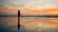 Silhouetted figure standing alone on wet beach sand during golden sunset with calm ocean waves and dramatic sky reflection.