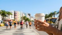 Hand holding disposable coffee cup in urban plaza with blurred crowd and city buildings in background, suitable for beverage, lifestyle, and urban commerce concepts.