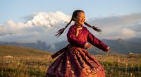 Young Asian woman in traditional burgundy dress dancing joyfully in mountain meadow at golden hour with snow-capped peaks and dramatic clouds in background.