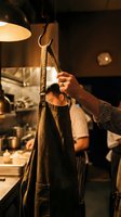 Chef hanging a worn canvas apron on a hook in a warm, busy restaurant kitchen with cooks working in the background.