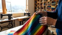 Senior craftsman hands holding vibrant rainbow striped textile in bright sewing workshop with vintage machine and fabric storage shelves.