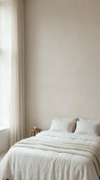 Minimalist bedroom interior with neutral beige walls, natural light from sheer curtains, and a neatly made bed with white linens and woven throw blanket. Suitable for home decor, interior design, and lifestyle content.