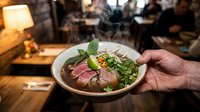 Asian noodle soup with sliced meat, fresh herbs, and vegetables served in bowl at restaurant with warm ambient lighting.