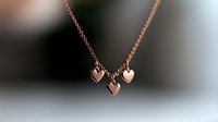 Delicate rose gold necklace with three small heart pendants on thin chain against blurred dark background, romantic jewelry accessory for special occasions.