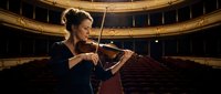 Professional female violinist performing solo in elegant concert hall with ornate golden architecture and red velvet seating during classical music recital.