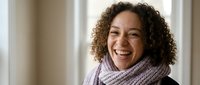 Happy mixed-race woman with curly hair wearing purple knit scarf smiling warmly indoors. Natural portrait expressing joy and positivity in cozy home setting.