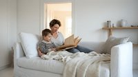 Mother and son reading book together on white bed in bright modern bedroom. Family bonding time, bedtime story, parenting moments at home.