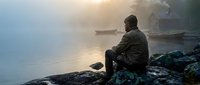 Man sitting on rocky shore contemplating misty lake at sunrise with boats and cabin in background creating peaceful morning atmosphere.