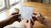 Senior adult hands holding blank white card on wooden table with natural window light, suitable for greeting cards, announcements, and promotional messaging.