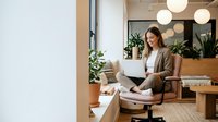 Young professional woman working on laptop in modern office space with plants and warm lighting for remote work and productivity concepts.
