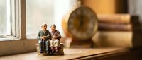Miniature elderly couple figurines sitting on bench in warm sunlit room with vintage clock and books creating cozy nostalgic atmosphere for home decor.