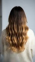 Young woman with long wavy ombre hair from behind wearing cream knit sweater against white wall for hair salon beauty and fashion concepts.