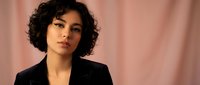 Professional young mixed-race businesswoman with curly hair in black blazer against pink backdrop for corporate branding and workplace diversity campaigns.