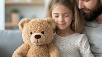 Caucasian girl with long blonde hair hugging soft brown teddy bear while sitting with bearded father on couch, expressing joy and tenderness in cozy home setting.