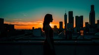 Silhouette of a woman standing on a rooftop overlooking a city skyline at sunset, golden hour lighting, urban landscape background, suitable for lifestyle and business concepts.
