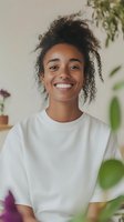 Young African American woman with natural curly hair and bright smile wearing white casual sweater against light background with decorative plants and flowers.