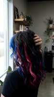 Woman with long black hair featuring vibrant blue and magenta highlights, shown from behind with hand touching hair. Modern interior with potted plants and wooden shelves visible. Suitable for beauty, hair care, and salon marketing.