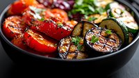 Grilled colorful vegetables in black bowl featuring charred tomatoes, eggplant and zucchini slices garnished with fresh herbs and balsamic glaze. Fresh summer meal.
