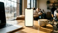Smartphone with blank white screen mockup on wooden table next to laptop and coffee cup in modern cafe workspace for app presentation and digital design.