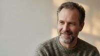 Smiling middle-aged Caucasian man with gray stubble wearing a knit sweater, looking away confidently against a light background.