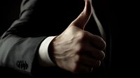 Close-up of businessman in formal suit showing thumbs up gesture against dark background, conveying approval, success, and positive feedback in corporate setting.
