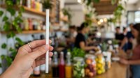 Unrecognizable person holding reusable bamboo straw in eco-friendly cafe with green plants and blurred customers, promoting sustainable living and zero waste lifestyle choices.