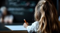 Young woman with blonde wavy hair sitting at desk writing in notebook, view from behind. Dark moody atmosphere with blurred background, focus on hair and shoulders.