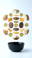 Floating burger ingredients with bread buns, cheese slices, meat patties and pickles falling into black bowl on white background for food design.