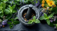 Stone mortar and pestle surrounded by fresh healing herbs, calendula flowers, and mint leaves on dark slate background, perfect for natural medicine and holistic wellness concepts.