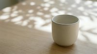 White ceramic cup on wooden table with dappled sunlight and shadows, minimalist design for home decor, lifestyle, and product photography.