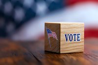 Wooden cube with American flag and vote text against blurred US flag background on rustic wooden surface, representing election concept.