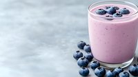 Fresh blueberry smoothie in glass with scattered berries on marble surface for healthy breakfast and nutrition concepts.