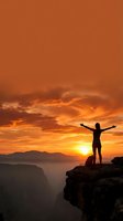 Silhouette of person with raised arms celebrating success on mountain cliff edge during dramatic orange sunset with canyon landscape below.