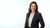 Professional businesswoman with long brown hair wearing black blazer and white shirt smiling confidently against clean white background for corporate use.