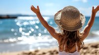 Young woman in straw hat with raised arms enjoying sunny beach day, view from behind against sparkling blue ocean water and bright summer sky.