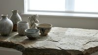 Handmade ceramic pottery collection with bowls and vases on rustic wooden table near window. Artisan crafted earthenware for home decor and kitchen use.