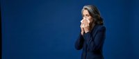 Middle-aged woman with shoulder-length brown hair and fair complexion, wearing a dark navy blazer, holding a tissue to her face against a solid blue background. Suitable for healthcare, illness, allergy, cold, flu, wellness and business-related content.
