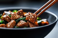Tender beef cubes in savory Asian sauce with stir-fried vegetables and sesame seeds served in black ceramic bowl, held with wooden chopsticks against dark background.
