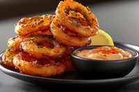 Crispy fried onion rings stacked on black plate with spicy dipping sauce and lemon slice, close up view of golden brown appetizer with herbs garnish.