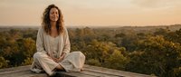 Young woman with curly hair meditating in lotus position on wooden deck at sunset with forest landscape background for wellness and mindfulness concepts.
