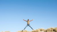 Shirtless young man jumping with arms raised against clear blue sky and dry grass field, expressing freedom and joy outdoors.