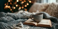 Steaming cup of coffee on open book with cozy knitted blanket and Christmas tree lights creating warm holiday atmosphere.