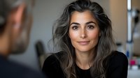 Young woman with long gray hair and natural makeup smiling warmly during conversation, wearing black sweater against blurred background, professional lifestyle portrait.