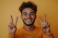 Young Hispanic man with curly hair showing peace signs and genuine smile wearing orange sweater against matching background. Positive emotional portrait.
