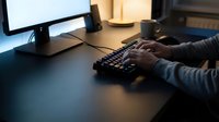 Professional working at computer desk typing on keyboard in modern office workspace with monitor and coffee cup during evening hours.