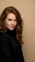 Confident middle-aged Caucasian woman with curly auburn hair wearing black turtleneck sweater against neutral beige background for professional headshot.