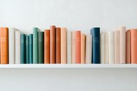 Row of hardcover books in pastel colors arranged on white shelf against light wall, creating minimal contemporary aesthetic for home library or bookstore display.